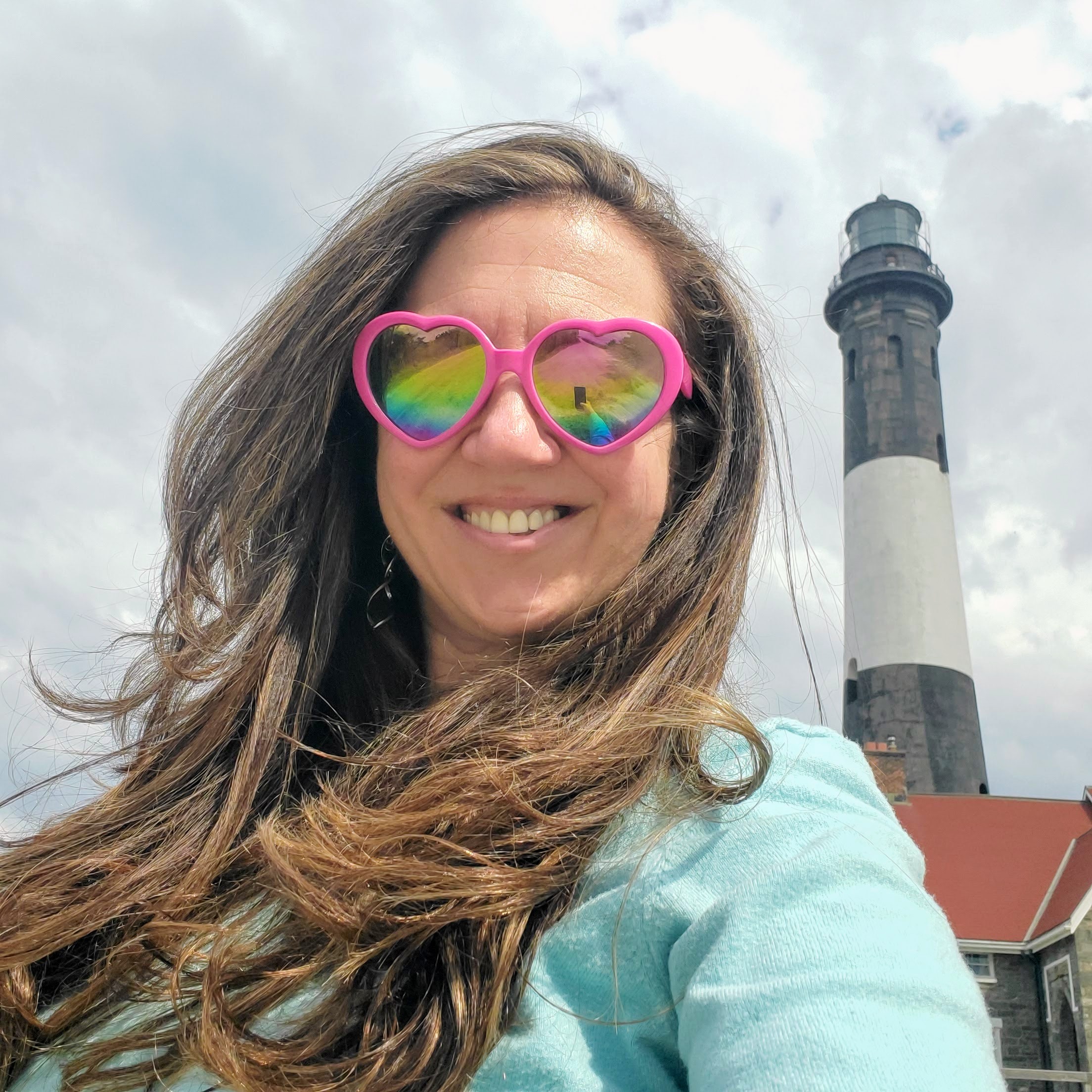 Natalie smiling outdoors near a lighthouse, wearing heart-shaped sunglasses